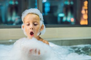 Happy little baby boy sitting in bath tub in the evening before going to sleep on the background of a window overlooking the evening city. Portrait of baby bathing in a bath full of foam near window